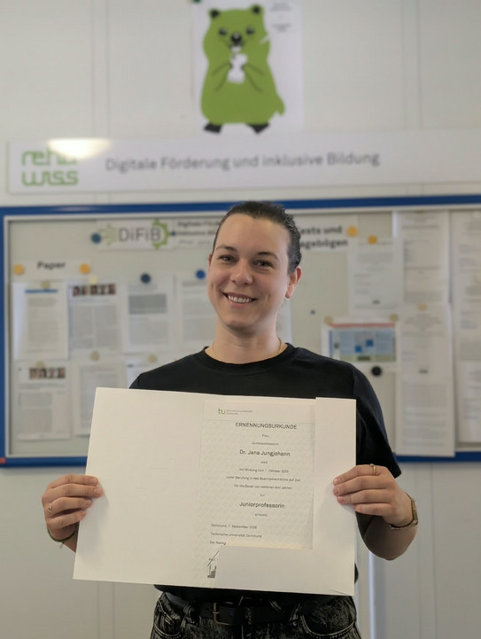 Jana Jungjohann steht vor der Informationstafel des Fachgebiets DiFiB und hält ihre Ernennungsurkunde für weitere drei Jahre als Juniorprofessorin in den Händen.