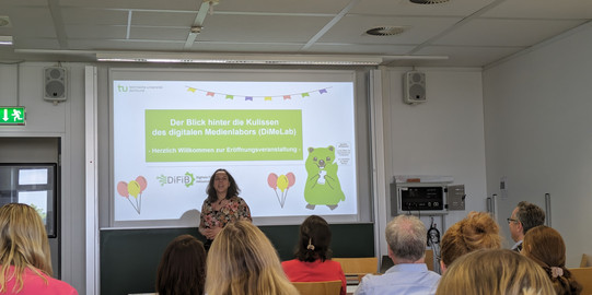 Foto von Jana Jungjohann vor ihrer Titelfolie zu Eröffnung des Digitalen Medienlabors. Auf der Titelfolie ist das Grußwort "Herzlichen Willkommen zur Eröffnungsveranstaltung" geschrieben.