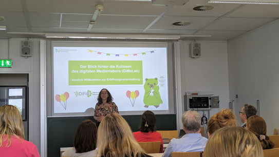 Foto von Jana Jungjohann vor ihrer Titelfolie zu Eröffnung des Digitalen Medienlabors. Auf der Titelfolie ist das Grußwort "Herzlichen Willkommen zur Eröffnungsveranstaltung" geschrieben. 