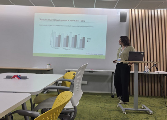 Foto von Jun.-Prof. Dr. Jana Jungjohann während ihres Symposiumsvortrags. Sie steht hinter einem Pult und zeigt auf die an die Wand projezierte Folie.
