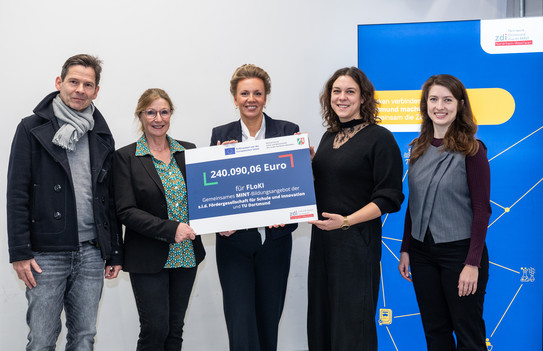 Fünf Personen stehen bei einer Förderbescheidübergabe in einem Innenraum. In der Mitte hält NRW-Wissenschaftsministerin Ina Brandes gemeinsam mit Vertreter*innen des Projektteams FLoKI einen großen Förderbescheid über 240.090,06 Euro.